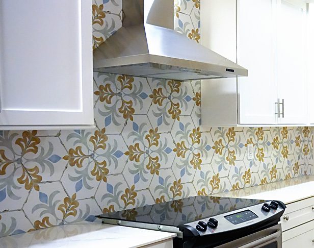Floral hex tiles with a white background and orange and light blue flowers on a kitchen backsplash with stainless steel range hood and white cabinets