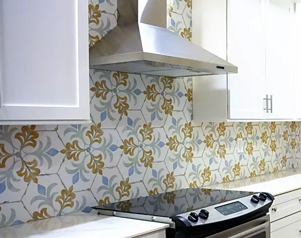 Floral hex tiles with a white background and orange and light blue flowers on a kitchen backsplash with stainless steel range hood and white cabinets
