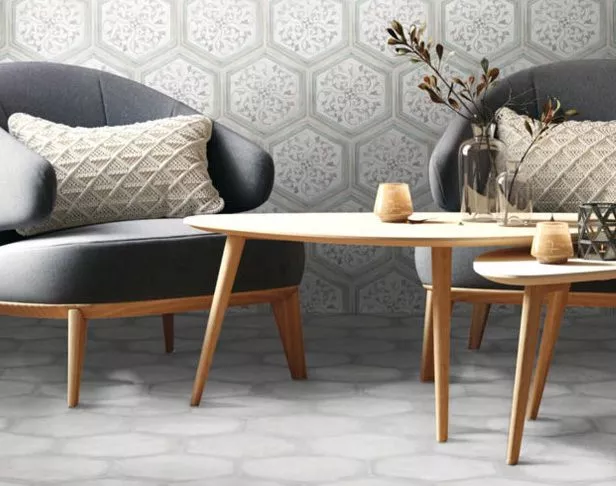 Hex wall tiles with a delicated floral pattern and white hex floor tiles in a sitting area with tow gray cushy chairs with pillows and accent tables