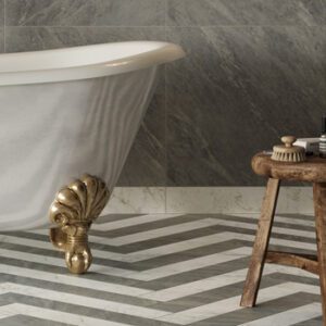 Gray and white marble look plank tiles on a bathroom floor in a chevron pattern with gold claw-foot tub and old wood stool