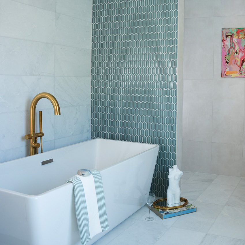 White marble-look tiled bathroom with acqua 3D mosaic tiles on one wall with large white soaking tub, gold faucet, and towels 
